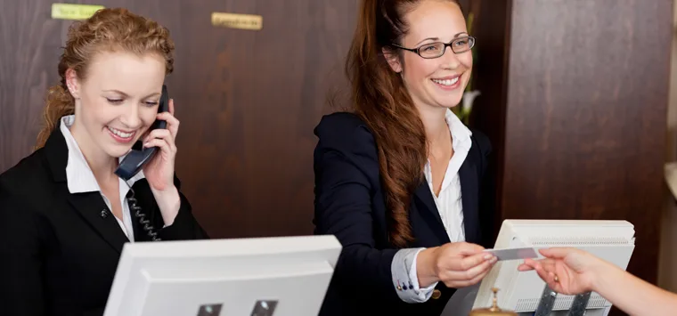 Mitarbeiterinnen an der Hotelrezeption telefonieren und bedienen einen Gast.